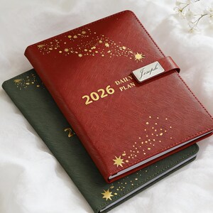 May include: Two leather-bound 2026 daily planners. The red planner has gold starburst designs and the word "Joseph" on a silver plate. The green planner is visible below. Both have gold lettering.