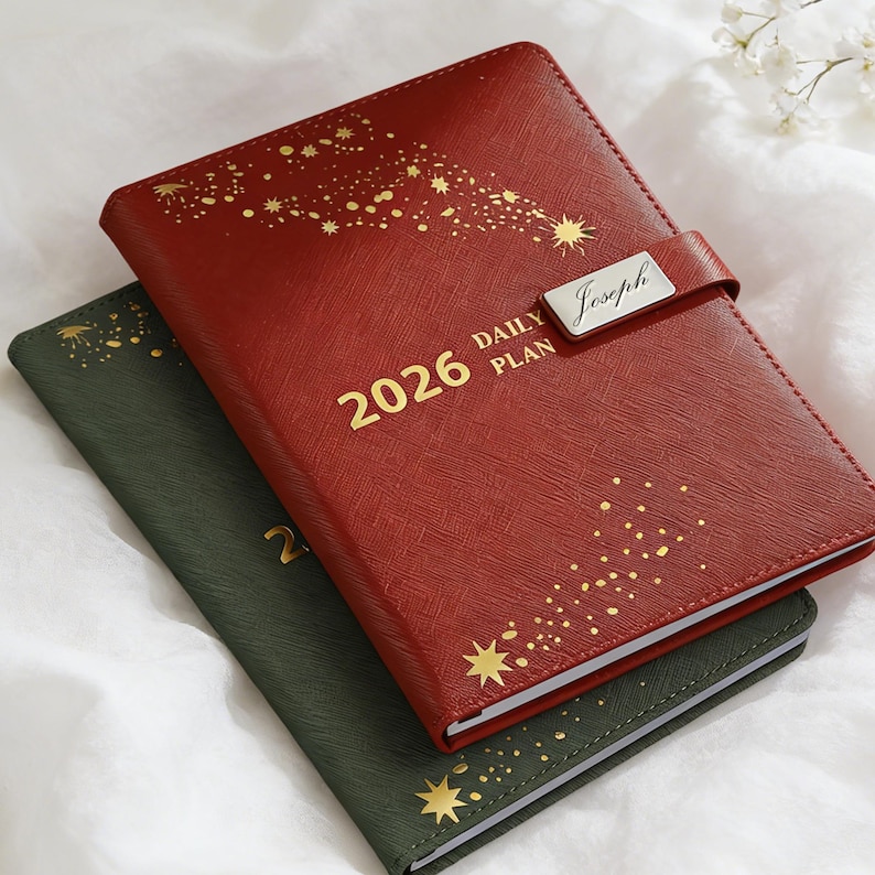 May include: Two leather-bound 2026 daily planners. The red planner has gold starburst designs and the word "Joseph" on a silver plate. The green planner is visible below. Both have gold lettering.