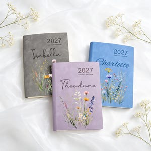 Może przedstawiać: Trzy kalendarze na rok 2027 w kolorach szarym, jasnoniebieskim i lawendowym. Każdy kalendarz posiada ilustrację polnych kwiat&oacute;w oraz spersonalizowane imię wydrukowane czarną kursywą: Isabella, Charlotte i Theodore. Okładki mają matowe wykończenie.