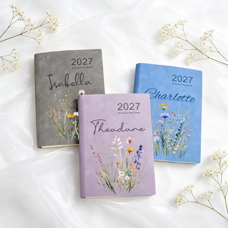 Może przedstawiać: Trzy kalendarze na rok 2027 w kolorach szarym, jasnoniebieskim i lawendowym. Każdy kalendarz posiada ilustrację polnych kwiat&oacute;w oraz spersonalizowane imię wydrukowane czarną kursywą: Isabella, Charlotte i Theodore. Okładki mają matowe wykończenie.