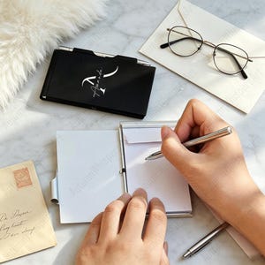 Op de afbeelding: Een zwart notitieblok met de naam "Amanda" in witte letters ligt op een marmeren oppervlak. Een persoon schrijft met een zilveren pen op een klein wit notitieblok. Een bril en een envelop liggen ook op het oppervlak.