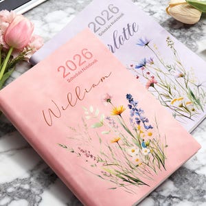 May include: Two 2026 schedule notebooks. One is pink with the name "William" and floral illustrations. The other is lavender with the name "Charlotte" and floral illustrations. Both have the year 2026 printed on the cover.