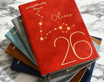 May include: A stack of weekly and monthly planners in various colors. The top planner is red with gold lettering that reads "weekly & monthly planner" and "Oliver". A constellation design and the number "26" are also visible.