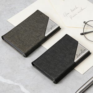 以下が含まれることがあります： 2つの名刺入れ。1つは濃いグレー、もう1つは黒で、シルバーのアクセントが付いています。濃いグレーのホルダーには「Sophia」という名前が刻まれ、黒いホルダーには「Amelia」と刻まれています。どちらも明るい灰色の表面にあります。