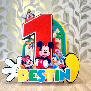 Puede incluir: Adorno de tarta de cumpleaños colorido con Mickey Mouse y sus amigos. El número "1" rojo es prominente, con el nombre "DESTIN" debajo. Incluye a Goofy, Pluto, Minnie Mouse, Donald Duck y Daisy Duck.