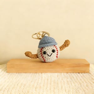 Puede incluir: Un llavero de béisbol de ganchillo con una cara sonriente, costuras rojas y una gorra azul. El béisbol tiene brazos marrones y un anillo de llavero dorado. El béisbol está sobre un bloque de madera.