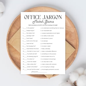 Puede incluir: Una tarjeta blanca de Office Jargon Match Game en una bandeja de madera. El juego presenta frases como "in the pipeline" y "the bottom line" con las definiciones correspondientes. También se ve un sobre marrón y acentos de algodón.