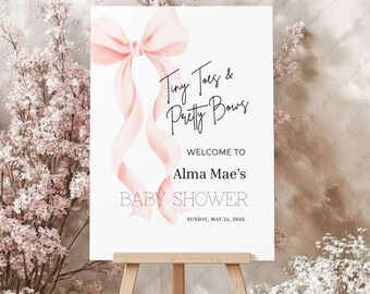 May include: A white baby shower sign on an easel, featuring a pink bow and ribbon design. The sign reads "Tiny Toes & Pretty Bows" and welcomes guests to Alma Mae's baby shower on May 24, 2026.