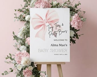 May include: A white rectangular sign on a wooden easel featuring a large watercolor-style light pink bow. The text reads: Tiny Toes & Pretty Bows, WELCOME TO, Alma Mae's, BABY SHOWER, SUNDAY, MAY 24, 2026. The sign is decorated with pink and white flowers.