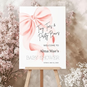 Puede incluir: Un letrero de baby shower sobre un caballete. El letrero presenta un gran lazo rosa y el texto "Tiny Toes & Pretty Bows". Debajo, se lee "Welcome to Alma Mae's BABY SHOWER Sunday, May 24, 2026."
