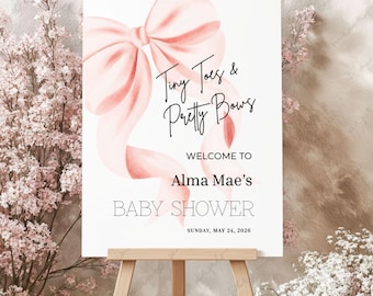 May include: A baby shower sign on an easel. The sign features a large pink bow and the text "Tiny Toes & Pretty Bows." Below, it reads "Welcome to Alma Mae's BABY SHOWER Sunday, May 24, 2026."