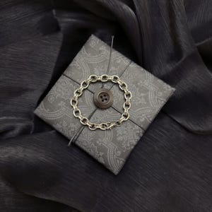 Pode incluir: Uma pulseira prateada com um design de elos de corrente, adornada com pequenas pedras transparentes, repousa sobre uma caixa de presente cinza escuro. A caixa é embrulhada com fio preto e apresenta um botão grande e escuro no centro.