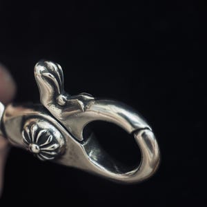 May include: Close-up of a silver-colored clasp with intricate floral and leaf-like details. The clasp has a curved, hook-like design and a decorative floral element. The background is a dark, solid color.
