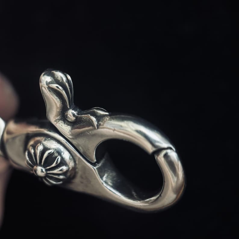 May include: Close-up of a silver-colored clasp with intricate floral and leaf-like details. The clasp has a curved, hook-like design and a decorative floral element. The background is a dark, solid color.