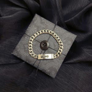 May include: A silver curb chain bracelet with a rectangular clasp featuring a cross design. The bracelet is displayed on a gray patterned gift box with a dark brown button. The background is a dark, textured fabric.