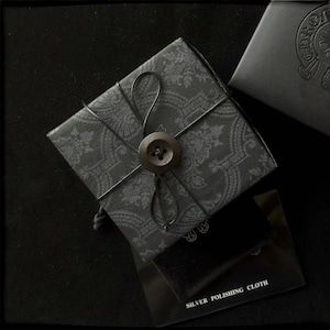 May include: A wrapped gift box with a paisley pattern, secured with black string and a large button. A small black box and a silver polishing cloth are also visible, all against a dark background.