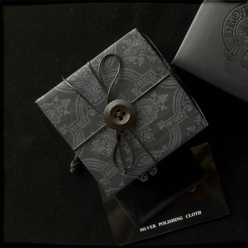 May include: A wrapped gift box with a paisley pattern, secured with black string and a large button. A small black box and a silver polishing cloth are also visible, all against a dark background.