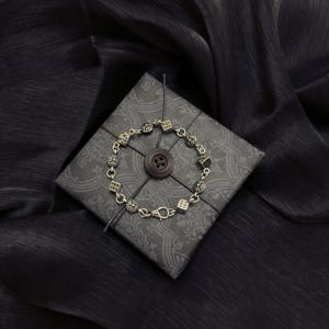 May include: Silver bracelet featuring dice and floral charms, displayed on a patterned grey gift box tied with black string. A large brown button is centred on the box. The background is a dark, textured fabric.
