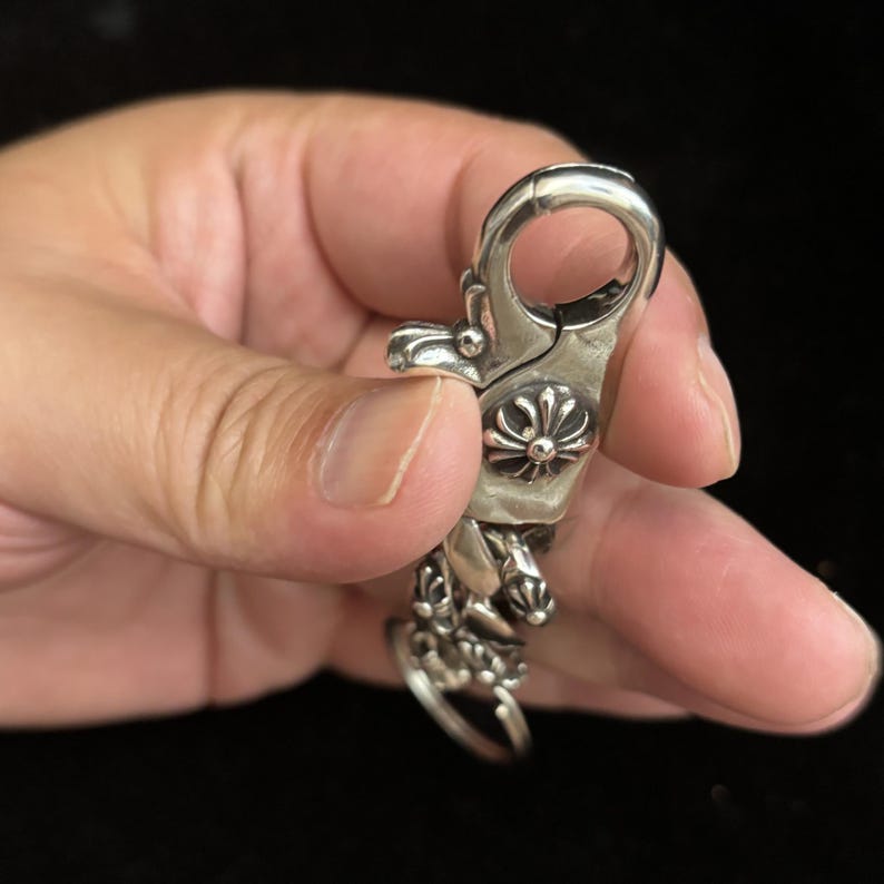 May include: A silver-colored metal clasp with ornate floral and geometric designs. The clasp has a large, rounded loop at the top and a decorative swivel base. The clasp is held in a hand against a dark background.