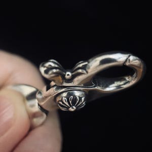 May include: Close-up of a silver-colored metal clasp with ornate floral and bow-like details. The clasp is held against a dark background, highlighting its intricate design and polished finish.