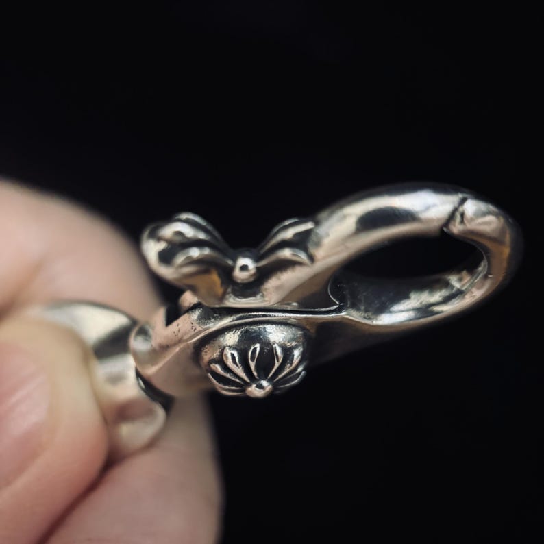May include: Close-up of a silver-colored metal clasp with ornate floral and bow-like details. The clasp is held against a dark background, highlighting its intricate design and polished finish.