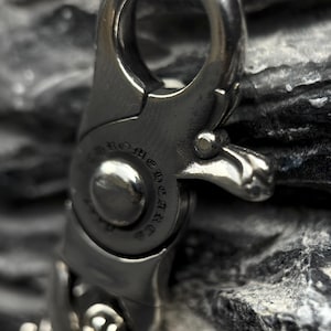 May include: Close-up of a silver-colored metal clasp with the engraved text "Chrome Hearts". The clasp has a hinged design with a lever for opening and closing. The background is a dark, textured surface.