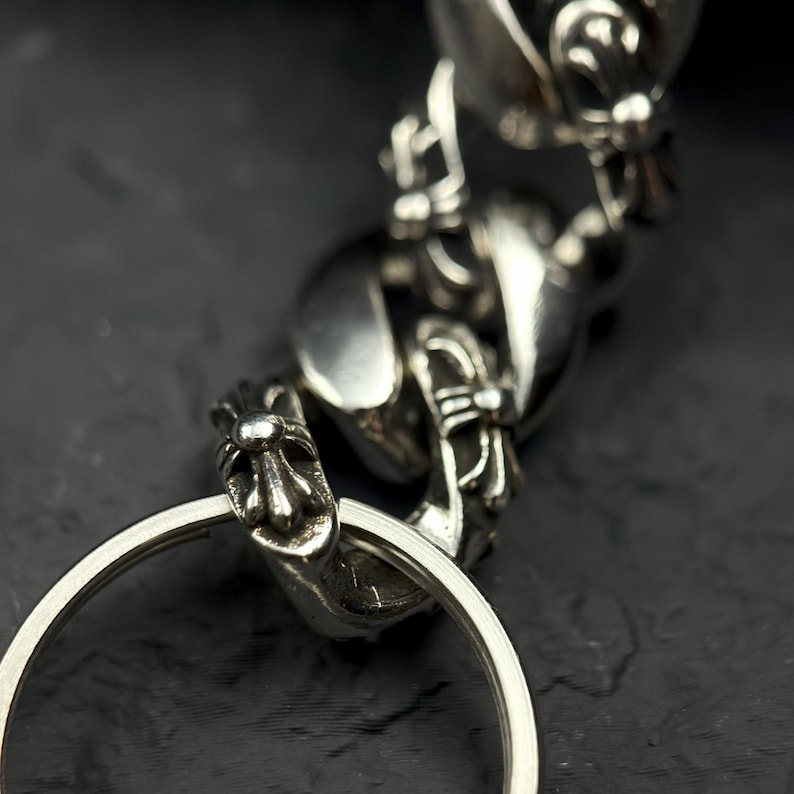 May include: Close-up of a silver-colored keychain with ornate, detailed links and a large, circular key ring. The design features a stylized, possibly gothic, aesthetic with decorative elements. The metal has a polished, reflective surface.