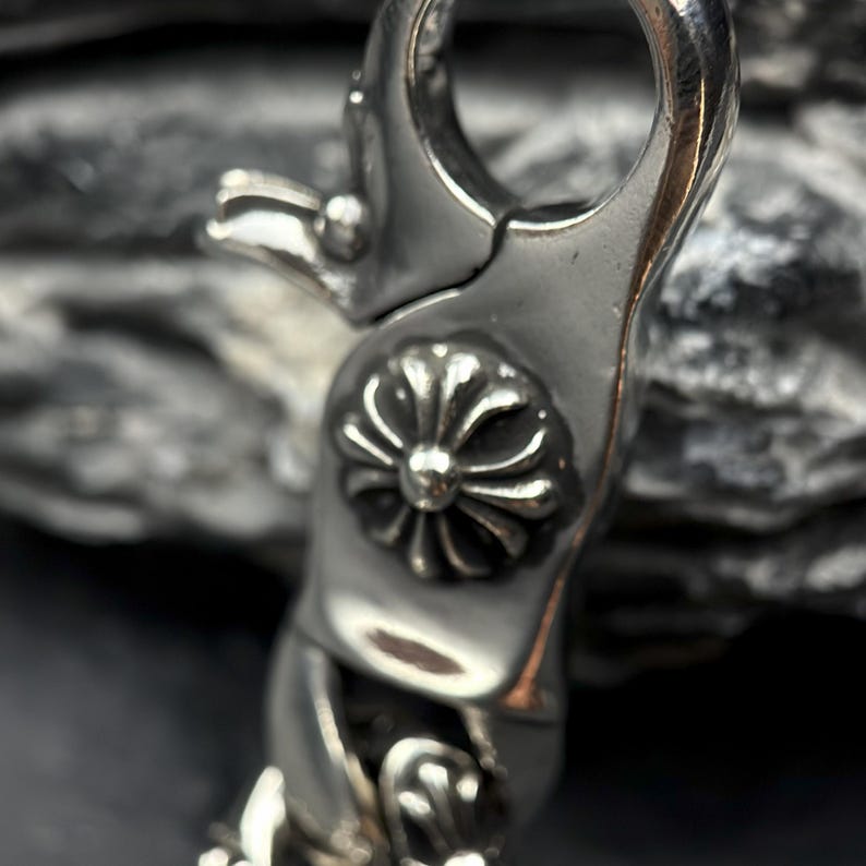 May include: Close-up of a silver-colored metal clasp. The clasp features a decorative floral design in the center and a curved, spring-loaded opening. The metal has a polished, reflective surface.