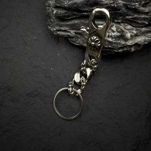 May include: Silver-toned metal keychain with a lobster clasp and a decorative floral design. The keychain features a chain link design and a circular key ring. The background is a dark, textured surface.
