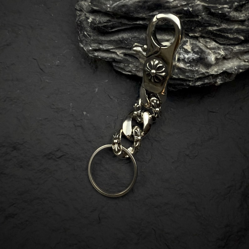 May include: Silver-toned metal keychain with a lobster clasp and a decorative floral design. The keychain features a chain link design and a circular key ring. The background is a dark, textured surface.