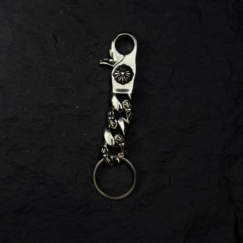 May include: A silver-colored key chain with a decorative floral design on the clasp. The chain is made of interlocking links and includes a key ring. The key chain is designed to attach to a belt loop or bag.