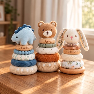 May include: Three stacking ring toys featuring plush animal toppers: a blue dinosaur labeled Lucas, a brown bear labeled Arlo, and a cream rabbit labeled Eloise. Each toy consists of various textured rings in shades of blue, green, brown, and cream, with some rings featuring star or floral patterns.