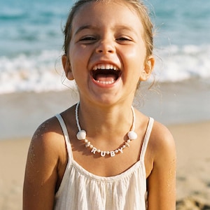 May include: A young person smiles broadly, wearing a white necklace with the name "RUBY" spelled out in white letter beads. The necklace also features white beads and seashell accents. The background shows a beach and ocean.