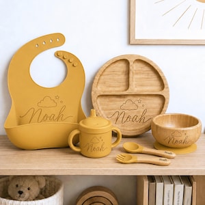 May include: A mustard yellow silicone bib, a mustard yellow sippy cup with two handles, a round three-section bamboo plate, a bamboo bowl with a yellow base, and a matching spoon and fork set. Each item is engraved with a cloud, stars, and the name Noah.