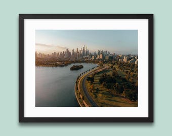 May include: An aerial photograph of a city skyline with a river and park in the foreground. The image is framed in a black border. The city has many tall buildings. The river curves around a small island. The park has trees and a road.