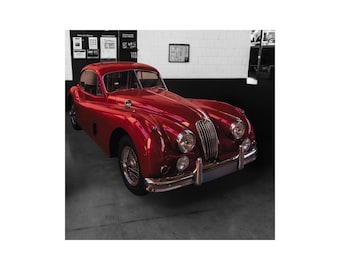 May include: A classic red vintage car with chrome accents, parked in a garage. The car has a sleek, aerodynamic design with rounded features, a long hood, and a prominent grille. The car's glossy finish reflects the surrounding environment.