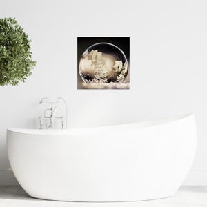 May include: A framed print of a clear bubble with intricate, crystallized patterns inside. The artwork is set against a dark background, with the bubble resting on a white, cloud-like base. The image is displayed on a white wall above a white bathtub.
