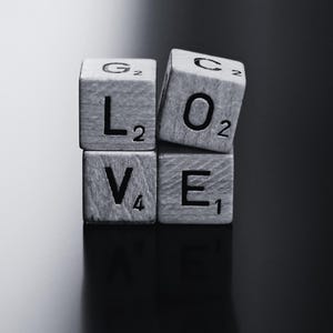 May include: A monochrome image showing wooden Scrabble tiles arranged to spell "LOVE". The tiles are stacked, with the letters and their point values visible. The background is a dark, reflective surface, creating a contrast with the tiles.