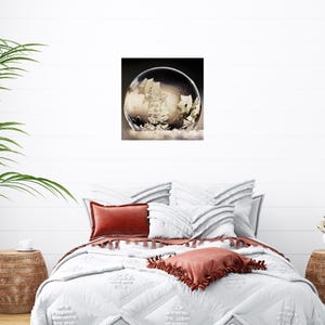 May include: A framed print of a clear bubble with a ship inside. The bubble is set against a dark background, with a white, cloud-like base. The print is displayed above a bed with white bedding and rust-colored pillows.