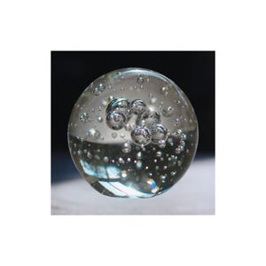 May include: A clear glass sphere with numerous air bubbles suspended within. The sphere has a cluster of larger bubbles at its center, and smaller bubbles throughout. The glass reflects light, creating highlights and a sense of depth. The overall effect is a delicate, almost ethereal appearance.