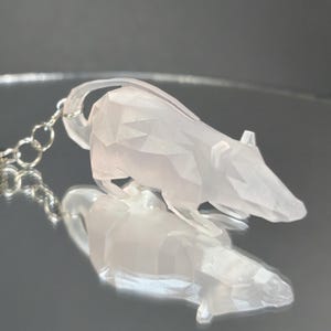 Könnte beinhalten: Ein durchsichtiger, weißer, geometrischer Ratten-Schlüsselanhänger mit einer silbernen Kette. Die Ratte ist aus einem glänzenden, facettierten Material gefertigt.