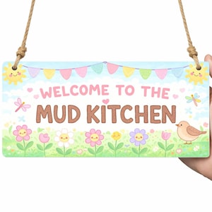 May include: A rectangular sign with the text "WELCOME TO THE MUD KITCHEN" in brown letters. The sign features a colourful design with flowers, a bird, and a banner. It is held by a person.