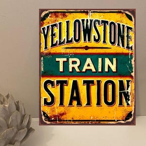 以下が含まれることがあります： レトロな雰囲気の長方形の金属看板。看板には「YELLOWSTONE TRAIN STATION」の文字が太字の白黒で、黄色、緑、赤の背景に表示されています。看板には茶色の縁取りがあります。