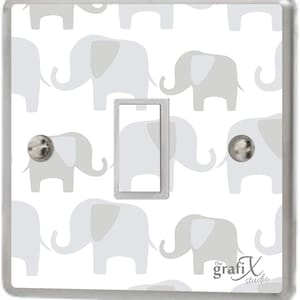 May include: A white light switch cover with a pattern of grey elephants. The switch cover has a silver frame and a clear plastic cover. The text "The grafi X studio" is printed in the bottom right corner.