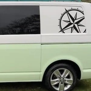 May include: A white van with a black compass sticker on the rear window. The compass features a black star at its centre and a circle of points surrounding it.