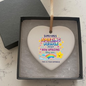 May include: A white heart-shaped ornament with the text "Sometimes Amazing People forget how amazing they are... This is your reminder." The ornament has a rainbow, stars, and cloud design. It is displayed in a black gift box with a gold ribbon.