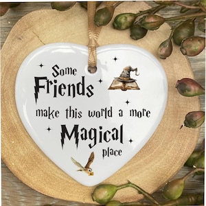 Könnte beinhalten: Weißer herzförmiger Keramik-Anhänger mit schwarzem Text, der "Some Friends make this world a more Magical place" lautet. Der Anhänger zeigt eine Schwarzweißillustration eines Zaubererhutes und eines Buches sowie eine weiße Eule mit braunen Flügeln.