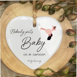 Peut inclure: Ornement blanc en forme de cœur avec le texte "Nobody puts Baby in a corner - dirty dancing." Un couple dansant est représenté au centre. L'ornement est suspendu à un ruban doré.