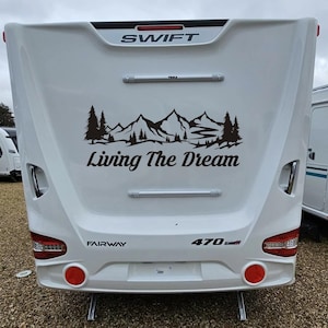 Può includere: Roulotte bianca Swift Fairway 470. Presenta una grafica nera di montagne e alberi con la scritta "Living The Dream". La roulotte ha luci posteriori rosse e una barra Thule. L'immagine è presa dalla parte posteriore.