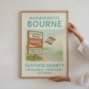 May include: A framed vintage-style poster for the Seafood Shanty in Bourne, Massachusetts. The poster features a sign with "Coca-Cola" and menu items, a bridge, and an "OPEN" flag. The poster is held by a person.